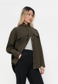 Femeie cu păr lung și blond, purtând o cămașă verde măsliniu, supradimensionată, peste un top gri deschis și pantaloni negri, cu mâna în buzunar.