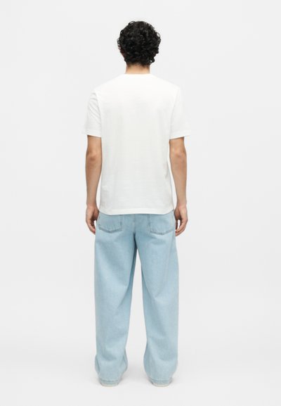 Άνδρας με σγουρά μαλλιά, φορώντας απλό λευκό μπλουζάκι και ανοιχτό μπλε τζιν παντελόνι με φαρδιά μπατζάκια, στέκεται με την πλάτη σε λευκό φόντο.