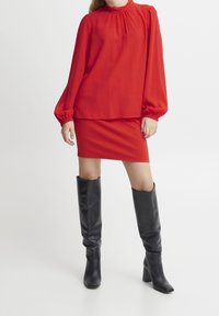 Rote Bluse mit langen Ärmeln und gerafftem Ausschnitt, kombiniert mit einem figurbetonten roten Rock. Schwarze kniehohe Stiefel mit Absatz, glatte Textur, minimales Design.