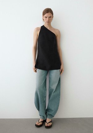 Femme portant un top noir asymétrique, un jean large bleu clair et des tongs plateformes noires, debout contre un mur blanc.