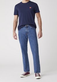 Camiseta de algodón azul marino con un pequeño logo rojo, combinada con vaqueros de mezclilla azul y zapatillas de lona color burdeos, sobre un fondo neutro.