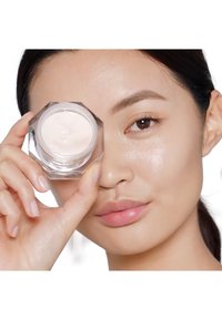 Clear glass jar containing a light cream. The jar has an angular design and is held by a person with light skin and natural makeup.