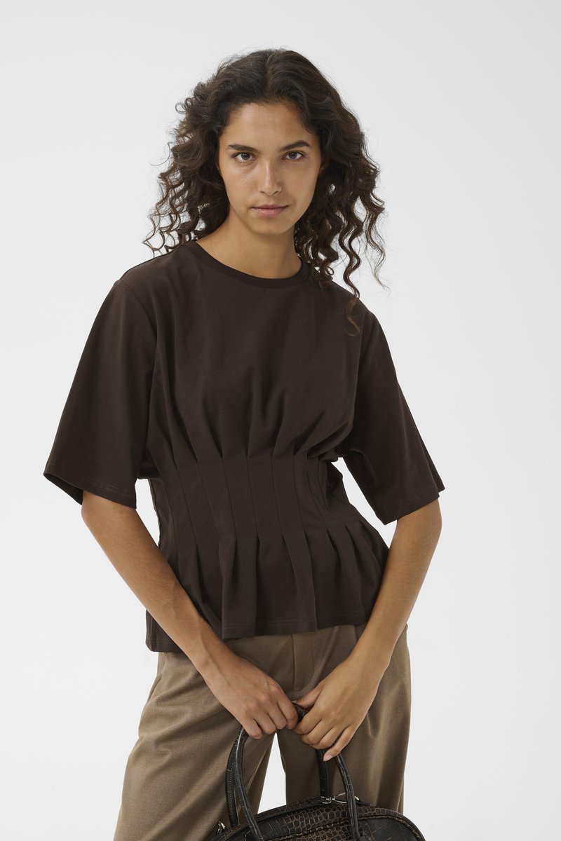Καφέ βαμβακερό μπλουζάκι με κοντά μανίκια και πτυχώσεις στη μέση, συνδυασμένο με μπεζ παντελόνι και μια υφασμάτινη μαύρη τσάντα.