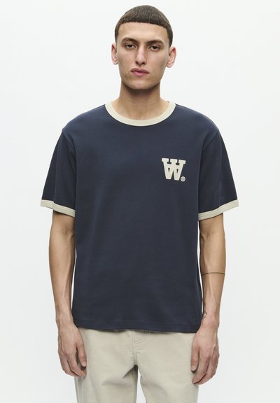 Ναυτικό μπλε βαμβακερό T-shirt με κρεμ ζακάρ γιακά και τονισμένα μανίκια. Διαθέτει λογότυπο στο στήθος σε αντίθετο ανοιχτό χρώμα.