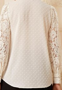 Bílá blůza s texturovanými puntíky a krajkovými rukávy. Dlouhé rukávy se zapínáním na knoflíky, mírně zakřivený lem a květinový krajkový vzor.