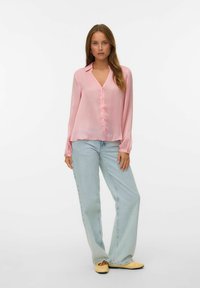 Chemise boutonnière rose clair à manches longues, encolure en V, texture douce, assortie à un jean taille haute bleu clair et des chaussures jaunes à enfiler.