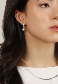 Kvinna med långt mörkt hår som bär en silverring med en hängande pärla och ett tunt silverhalsband på en vit topp.