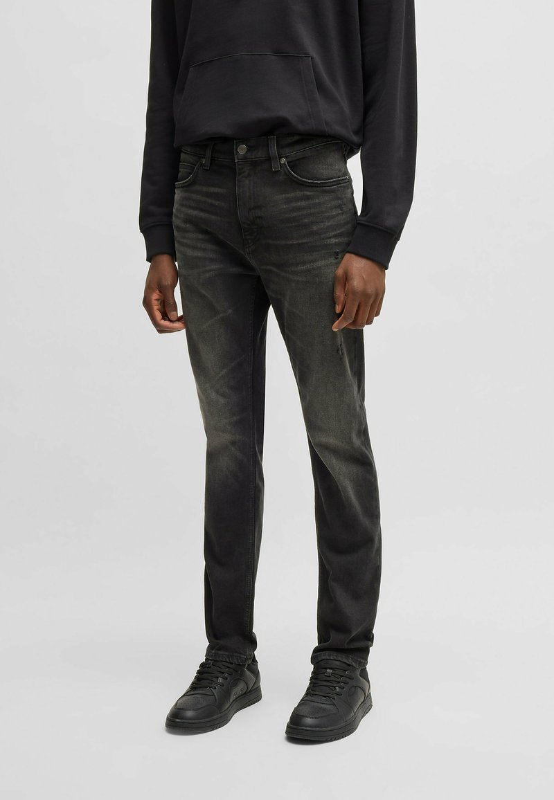 Jeans in denim nero con una vestibilità slim, caratterizzati da un lavaggio scolorito, dettagli cuciti, cinque tasche e una texture usurata, abbinati a sneakers nere.