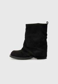 Botas de tornozelo em camurça preta com um design deslizante, bico arredondado e sola plana de borracha. Apresentam uma alça na parte de trás para facilitar o calçar.