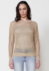 Beige, kötött pulóver laza hálós dizájnnal, hosszú ujjal és kerek nyakkivágással, világoskék farmernadrággal párosítva.