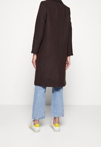 Hnědý kabát ze směsi vlny s límcem, dlouhými rukávy a rovný střih, nošený přes světle modré široké džíny a bílé tenisky s žlutými akcenty.