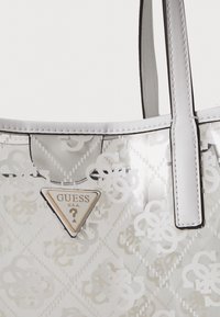 Sac cabas blanc avec une finition brillante, un motif de logo entrelacé et une étiquette triangulaire en métal avec logo à l'avant. Texture lisse, forme structurée.