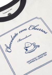 Chemise blanche avec col noir comportant un texte bleu "Chocolate con Churros Barcelona" et une tasse avec un motif de crème fouettée.