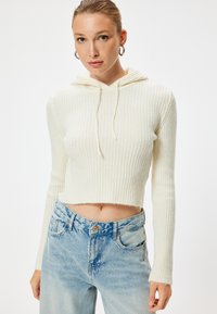 Ausgeschnittene weiße Ripp-Hoodie mit Kordelzug, langen Ärmeln und weicher Textur, kombiniert mit hellblauen, destruierten, hoch taillierten Jeans.