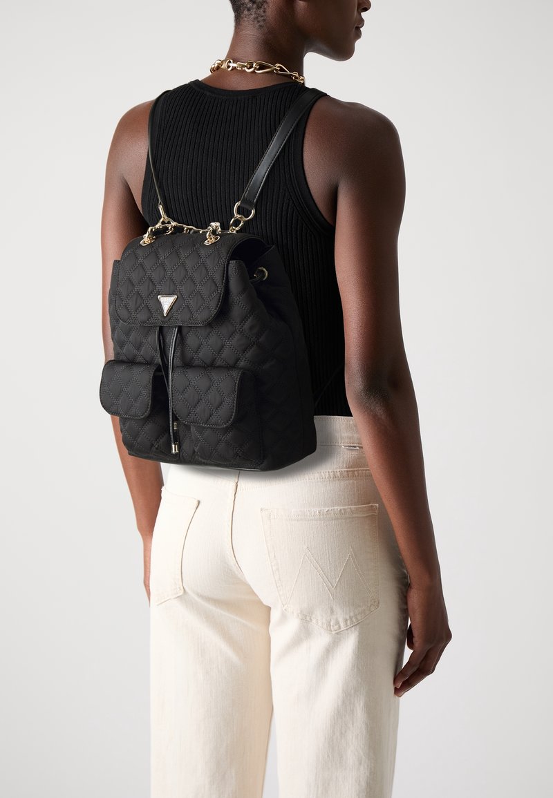 Personne portant un sac à dos matelassé noir avec des détails en chaîne dorée, un débardeur noir, un pantalon blanc et un collier en chaîne dorée, vue de dos.