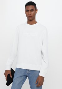 White sweatshirt with crew neck and embossed "MICHAEL KORS" logo on the front, paired with blue denim jeans and holding a black clutch.