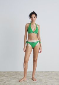 Set di bikini verde con top a collo halter e slip con lacci laterali. Entrambi i pezzi presentano il logo "Tommy Hilfiger" in bianco sulla banda.