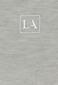 Hellgrauer Stoff mit weicher Textur, auf dem ein weißes, gesticktes "LA"-Logo innerhalb eines Quadrats präsentiert wird. Standardausführung mit Rundhalsausschnitt.