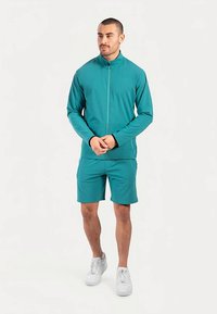 Teal athletic jacket with a high collar, paired with matching shorts. Both made of smooth, stretch fabric; white sneakers complete the outfit.