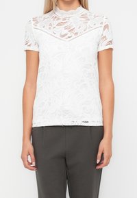Top corto bianco in pizzo con motivi floreali indossato con pantaloni grigio scuro, concentrandosi sulla parte centrale e sulle braccia su uno sfondo semplice.