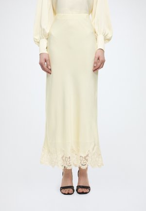 Kobieta w kremowej spódnicy midi z atłasu z koronkową lamówką na dole, w połączeniu z kremową bluzką z długim rękawem i czarnymi sandałami na otwartym palcu.