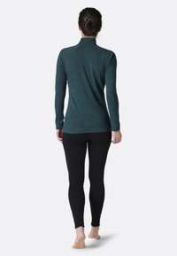 Long-sleeve top in dark teal, made of textured fabric with a high collar. Paired with black leggings, showcasing a fitted silhouette.