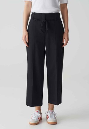 Black wide-leg trousers with a drawstring waist and front pleats, made from smooth fabric. Paired with white sneakers featuring red accents.
