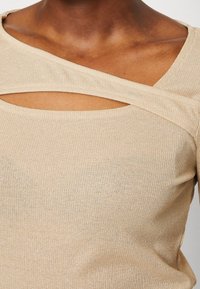 Top beige à côtelé présentant un design unique avec des découpes et des bretelles croisées. Texture douce avec une légère finition scintillante.