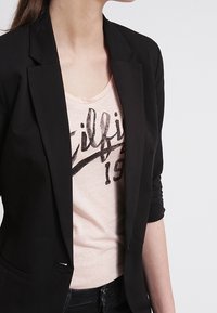 Černý blazer s přiléhavým střihem, zapínání na jeden knoflík a hladkou texturou, vrstvený přes světle růžové tričko s grafickým textem.