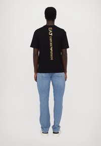 Camiseta negra de algodón con texto vertical amarillo "EA EMPORIO ARMANI" en la parte posterior. El modelo lleva jeans de mezclilla azul claros y zapatillas blancas.
