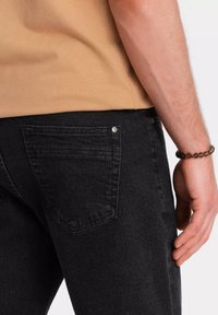Los jeans de mezclilla oscuros tienen un ajuste slim, bolsillos traseros con detalles de costura y un brillo sutil, combinados con una camiseta de cuello redondo beige.