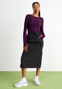 Langärmliges lila Oberteil mit einem spiralen Muster, kombiniert mit einem schwarzen Midirock mit Taschen und Kordelzug. Abgerundet mit Sneakers.