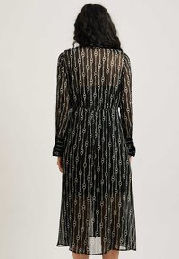 Abito nero trasparente con un motivo a catena, dotato di maniche lunghe, vita cinchata e parte inferiore fluente che arriva a metà polpaccio.