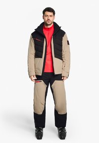 Ensemble veste et pantalon de ski en beige, noir et rouge. La veste présente un design matelassé avec des zips ; le pantalon possède des poches latérales et des sections en coloris contrastés.