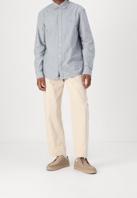 Knappad skjorta i ljusblått tyg, beige avsmalnande byxor och bruna sneakers med texturerade accenter. Märkbart logo på skjortans bröst.