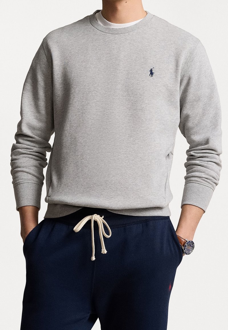Homme portant un sweat-shirt gris clair à col rond avec un petit logo bleu marine et un pantalon de survêtement bleu marine avec un cordon blanc, mains dans les poches et montre au poignet gauche.