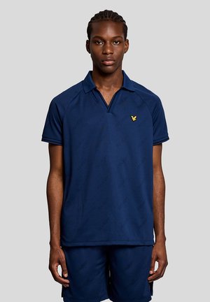 Jonge man met gevlochten haar, gekleed in een donkerblauw poloshirt en bijpassende korte broek, staand tegen een effen lichtgrijze achtergrond.