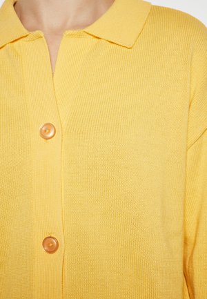 Cardigan tricotat galben, cu un guler clasic, închidere cu nasturi și un material texturat. Accentuate cu un nasture rotund, de culoare deschisă.