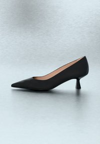 Escarpin noir à bout pointu et talon bas avec une finition lisse sur une surface grise réfléchissante.