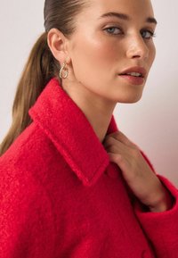 Rød uldfrakke med tekstureret finish og hakket krave. Modellen bærer gyldne øreringe i hoop-stil og rører ved kraven med den ene hånd.