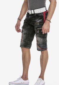 Zwarte denim shorts met een vervaagde afwerking, voorzien van meerdere met rits gesloten zakken, rode accenten en een witte gesp met studs. Witte sneakers maken de look compleet.