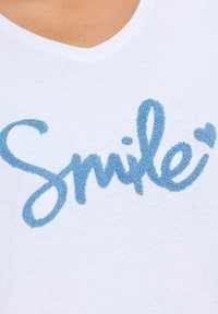 Bílý bavlněný tričko s výstřihem do V, které má texturované modré písmo "Smile" se srdcovým akcentem, vyrobené z lehké a měkké tkaniny.