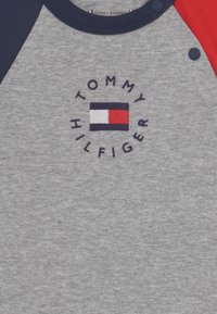 Szary bawełnowy t-shirt z granatowymi i czerwonymi rękawami raglanowymi. Zawiera haftowane logo "TOMMY HILFIGER" oraz prostokątną naszywkę z logo na środku.