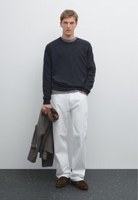 Marineblauer Pullover, graues T-Shirt darunter, weiße Hose. Hält einen braunen Mantel, trägt braune Wildlederschuhe. Einfaches Design, minimalistische Texturen.