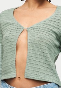 Γυναίκα που φοράει ένα cropped ζακέτα πλεκτή σε ανοιχτό πράσινο με υφή, κουμπωμένη με ένα κουμπί, που αφήνει εμφανές το μέσο της κοιλιάς πάνω από ανοιχτόχρωμο τζιν παντελόνι.