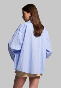 Light blue and white striped oversized shirt with dropped shoulders, mid-length sleeves, and a curved hem. Smooth cotton fabric.