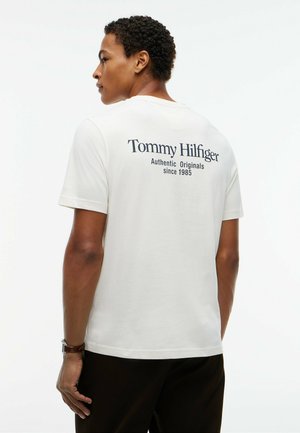 Hombre con cabello corto y rizado, que lleva una camiseta blanca de Tommy Hilfiger con texto en la espalda "Authentic Originals since 1985" y pantalones negros.