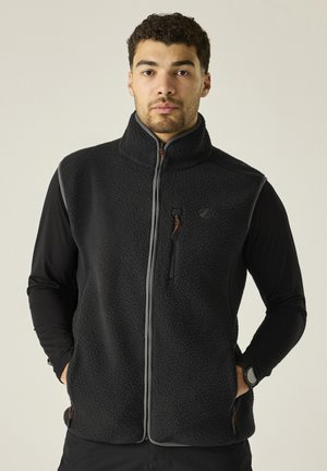 Young man wearing a black fleece vest over a black long-sleeve shirt, standing with hands in vest pockets, neutral background.