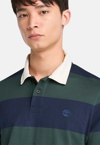 Camisa polo con franjas horizontales en verde oscuro y marino, un cuello color crema y un pequeño logo bordado en el pecho.