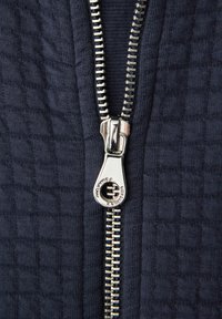 Nærbillede af sølvfarvet metallynlåstrækker på mørkeblå tekstureret stof, lynlåsen er delvist åben og viser sammenlåste tænder langs kanterne.